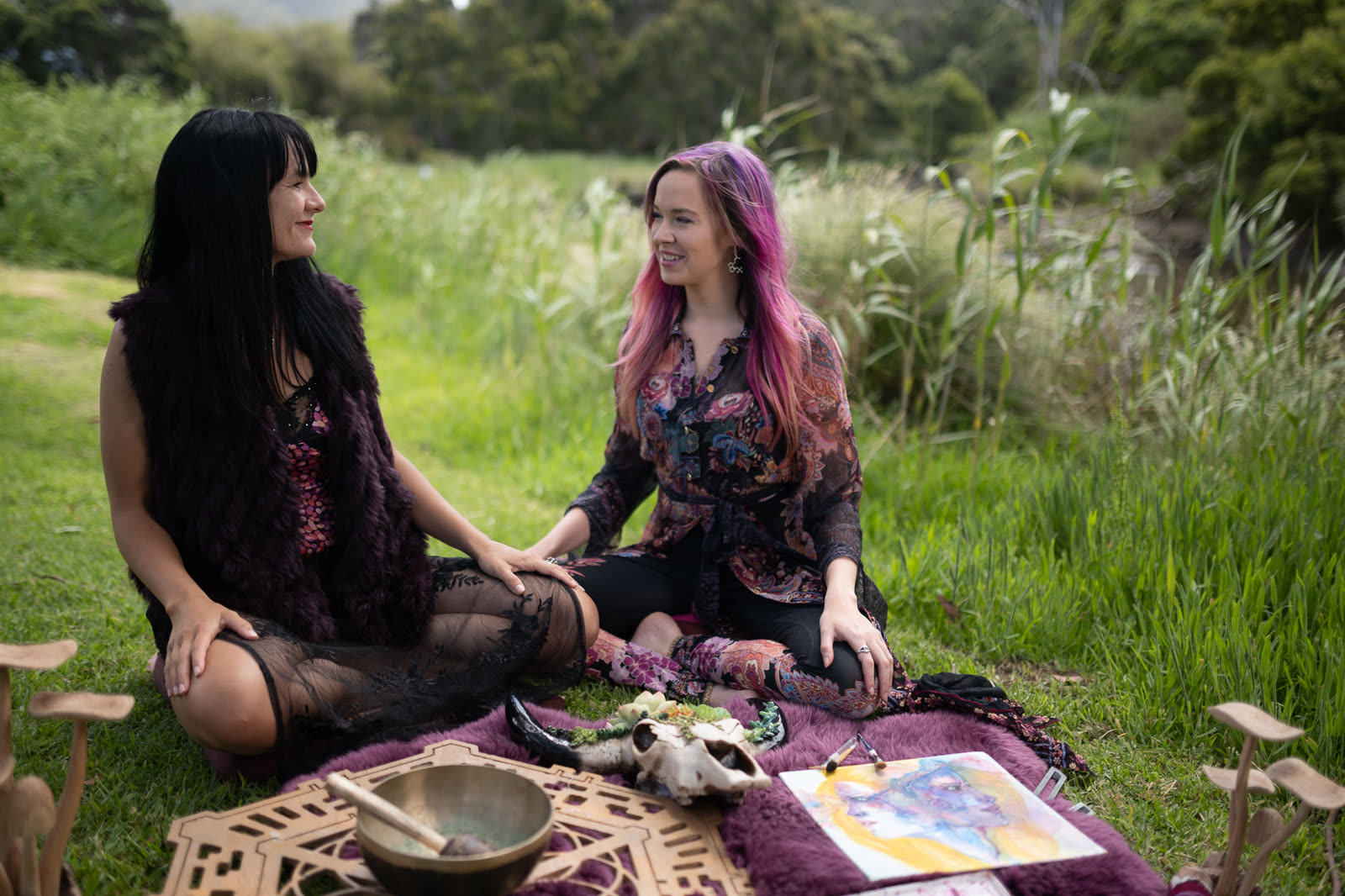 Dr Lani Roy, with long dark hair, and Melissa Warner, with pink hair, sitting together on a picnic rug in a green landscape, hands joined and turned toward one another